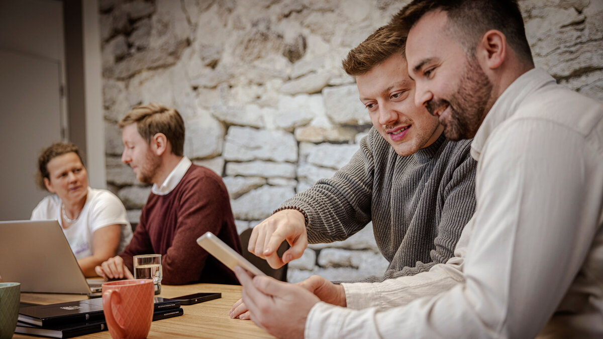Vier gutgelaunte Mitarbeitende sitzen an einem Konferenztisch und arbeiten gemeinsam an einem Laptop und Tablet.
