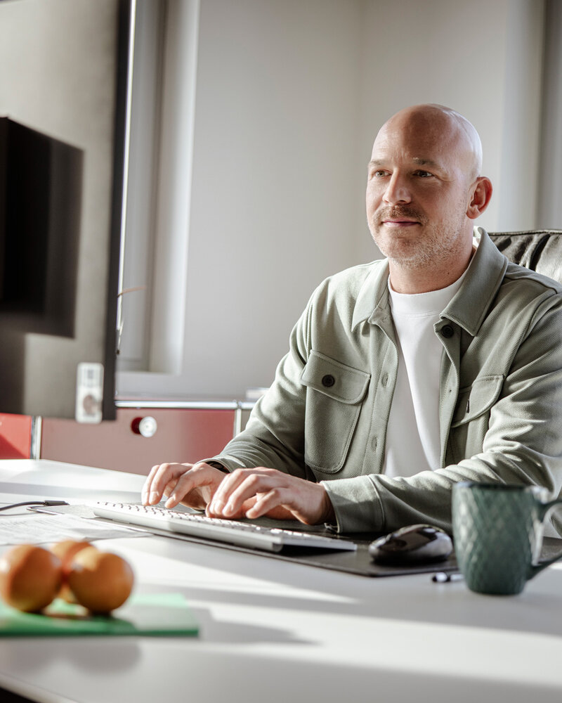 Mitarbeiter, der an seinem Schreibtisch sitzt, sympathisch lächelt und auf seinen Monitor schaut.